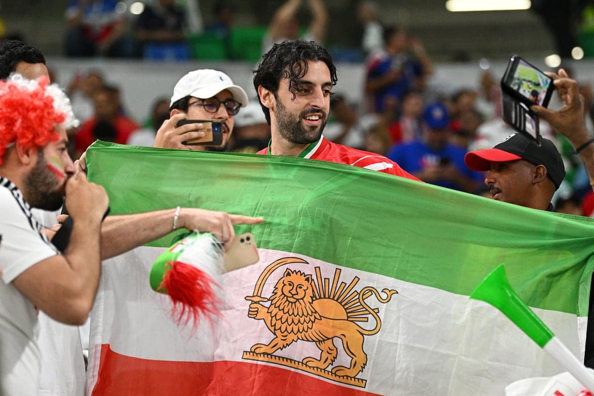 A stylized rendering of the pre-revolutionary Iranian flag with a lion and sun motif, set against the backdrop of a packed World Cup stadium.