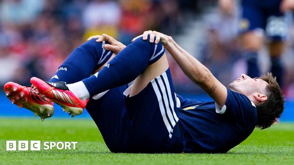 Billy Gilmour sitting on the Hampden Park turf clutching his knee while being comforted by Scotland teammates during a friendly match.