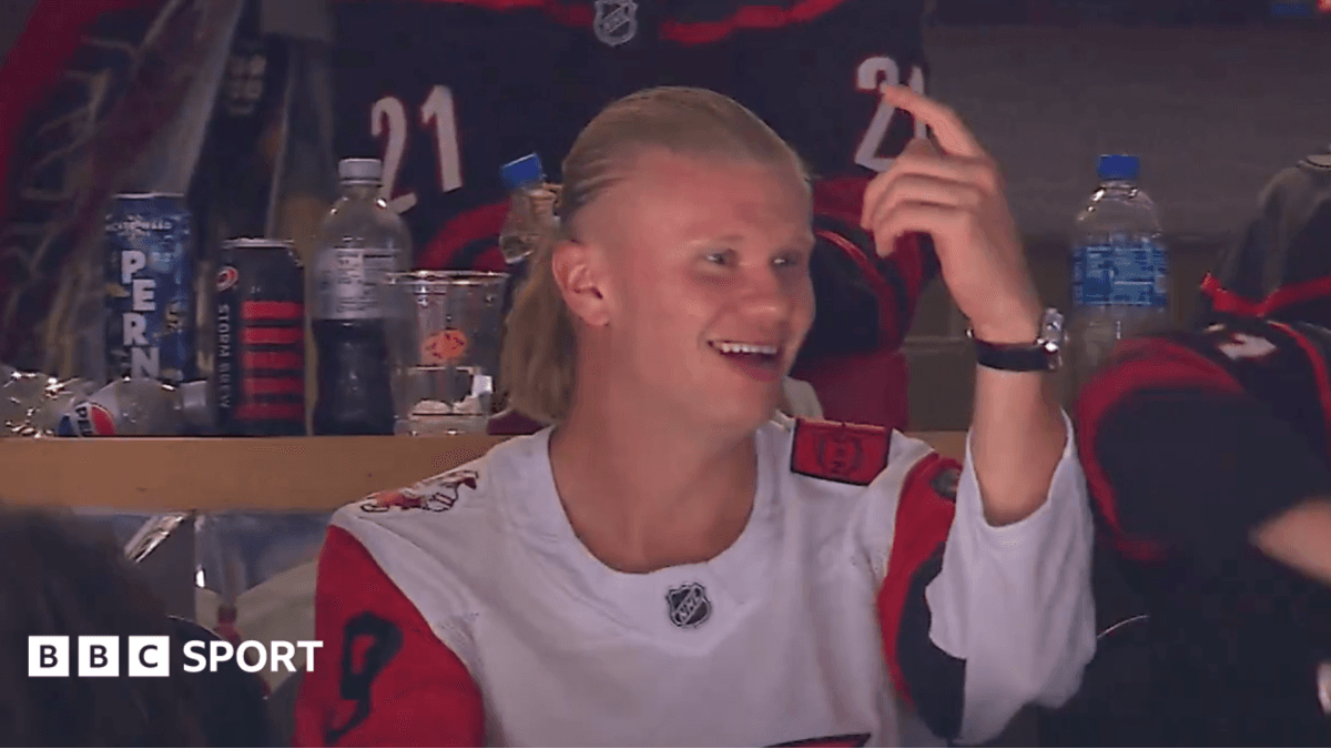 Erling Haaland in a white Carolina Hurricanes jersey with red and black sleeves, smiling and pointing his left hand upward while attending a Stanley Cup Final game.