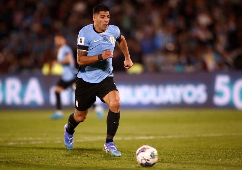 Luis Suarez looking dejected on the pitch in a Uruguay jersey, with coach Marcelo Bielsa visible in the background shouting instructions.