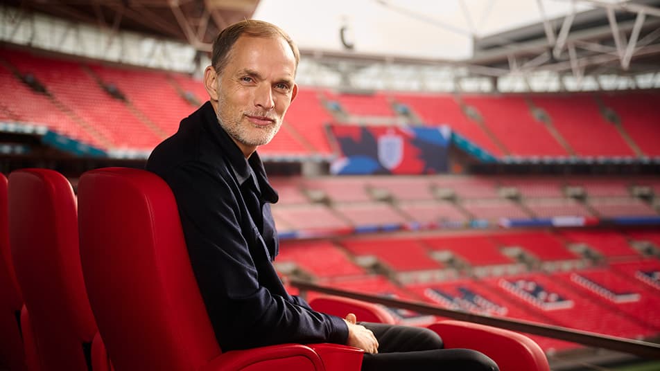 Thomas Tuchel looking pensive on the touchline, with Phil Foden and Cole Palmer pictured in the blurred background during a Premier League match.